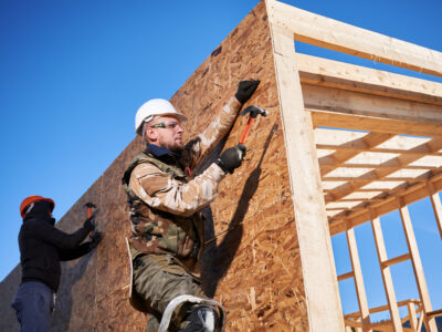 Carpenters hammering nail into OSB panel on the wall of a house.