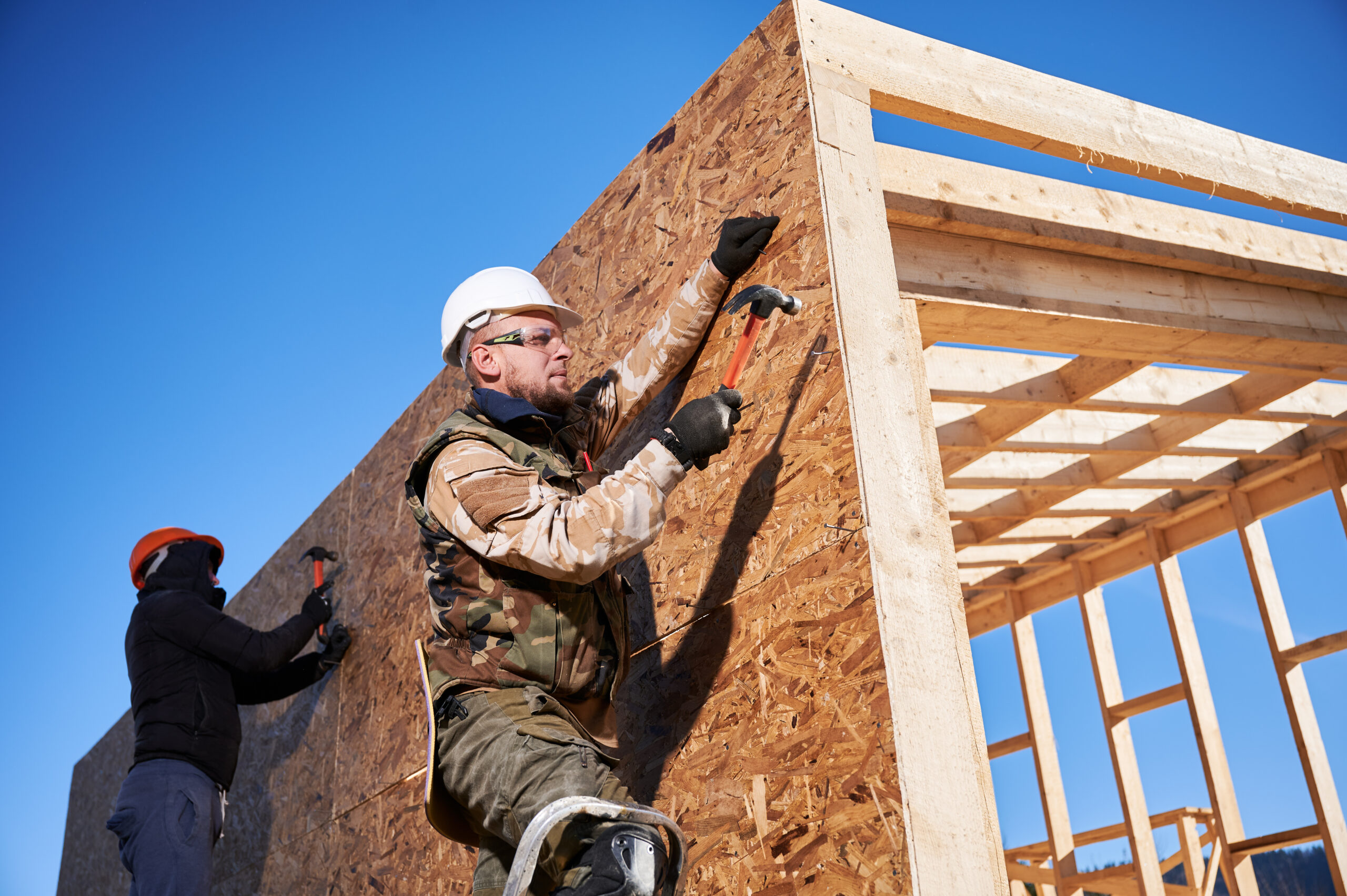 Carpenters hammering nail into OSB panel on the wall of a house.