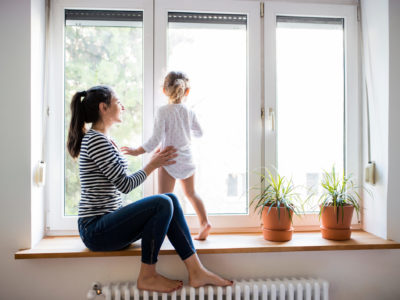 Mother sits on a window sill with child standing on hot water radiator
